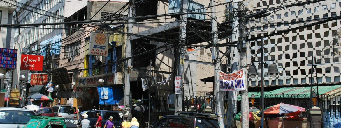 Busy street scene with tangled overhead wires, multiple signs, cars, and pedestrians in front of old buildings. The area appears crowded, with dense urban structures and various storefronts visible.