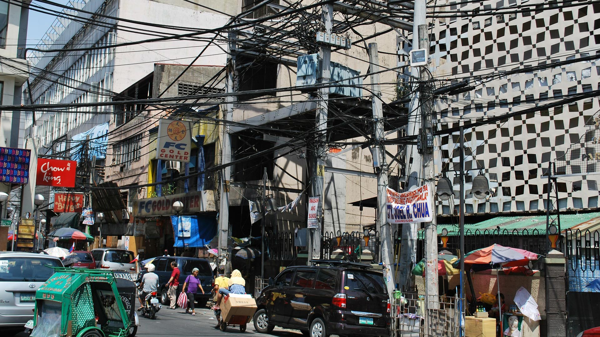 Busy urban street scene with tangled overhead wires, cars, pedestrians, street vendors, and buildings in the background. Signs and banners hang on storefronts, creating a crowded and lively atmosphere.