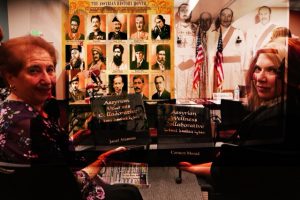 Two women sit smiling in front of a display featuring historical Assyrian figures and American flags. They each hold books titled Assyrian Wellness Collaborative. Posters and portraits fill the background.