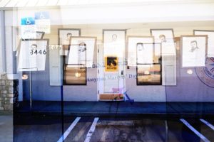Framed portraits and documents are displayed in a window, reflecting a building with a RESCUE sign, parking spaces, and a door decorated with Assyrian Martyrs Day signage.