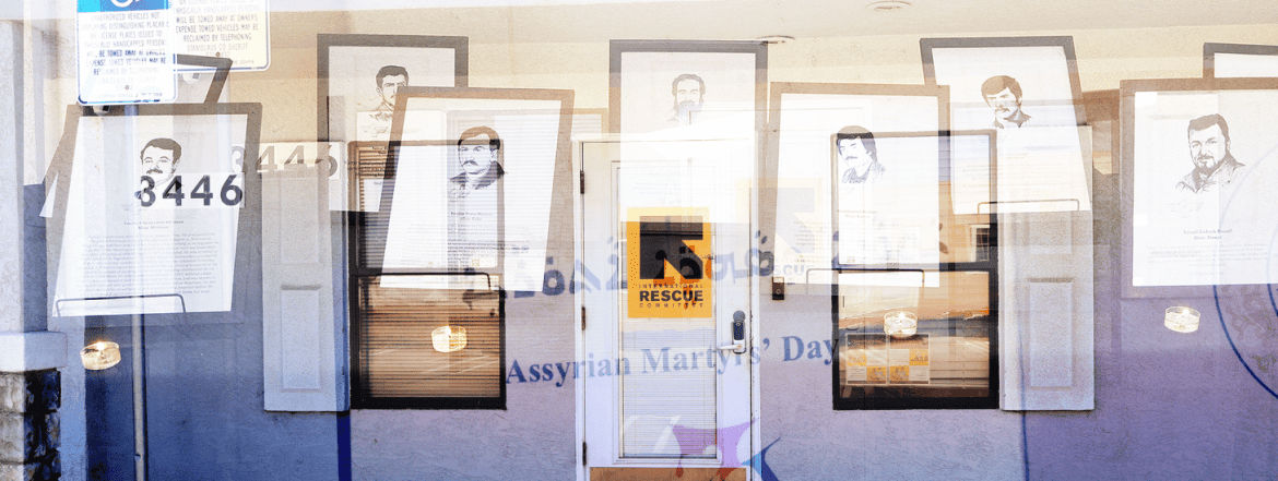 A storefront with a white door and the number 3446 displays Rescue signage. Several portraits of men with mustaches are displayed, along with text titled Assyrian Martyrs Day.
