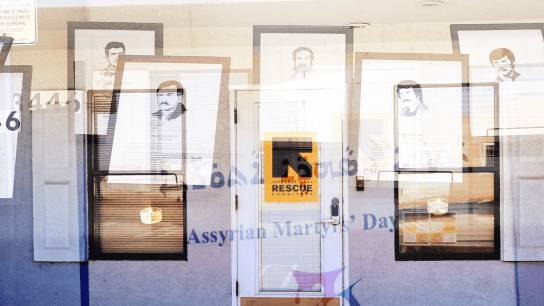 A storefront with a white door and the number 3446 displays Rescue signage. Several portraits of men with mustaches are displayed, along with text titled Assyrian Martyrs Day.